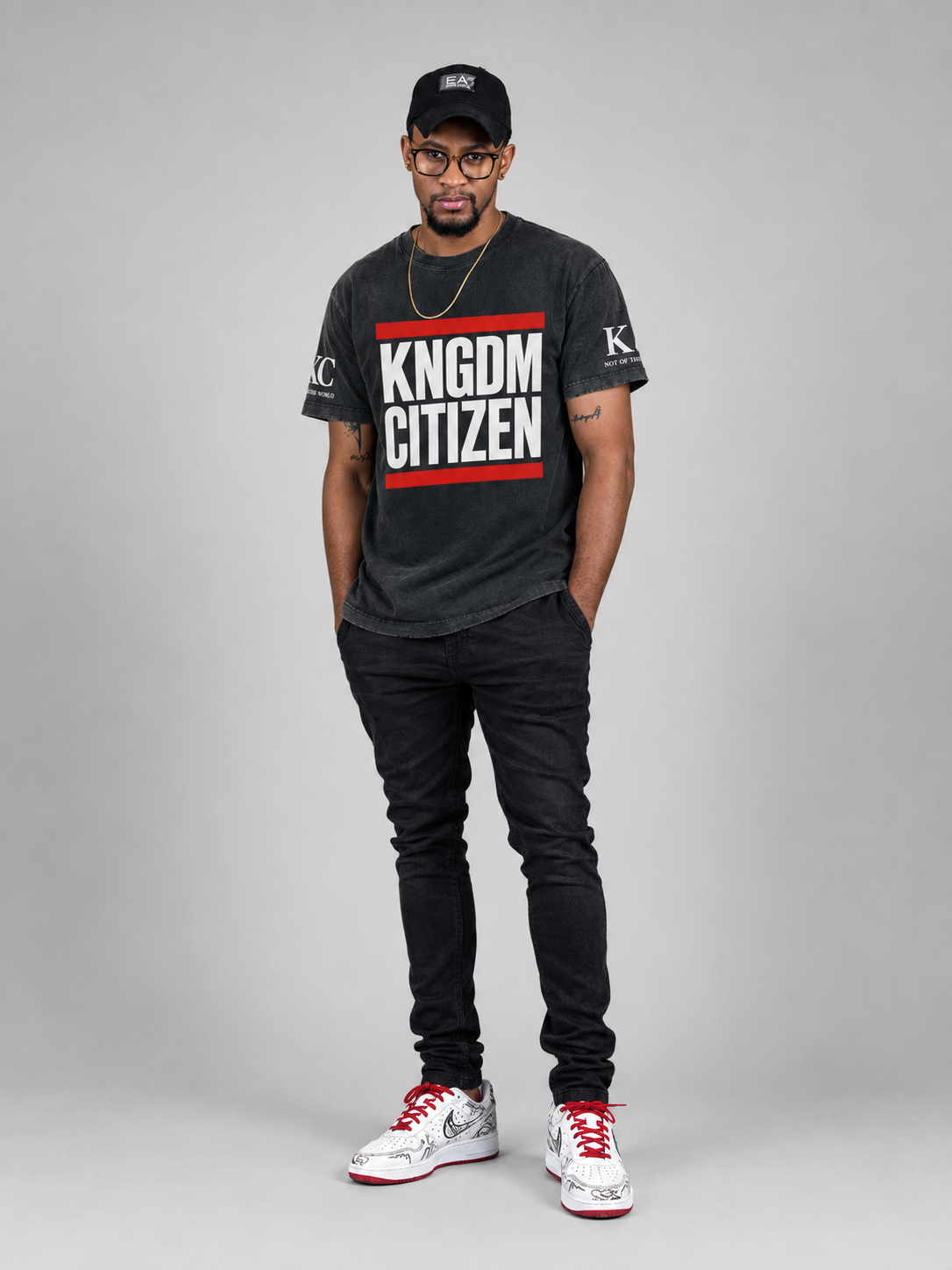 A man modeling a black graphic t-shirt with 'KNGDM CITIZEN' printed in white and red, paired with black jeans, a black cap, and white sneakers with red accents, against a solid gray studio background.
